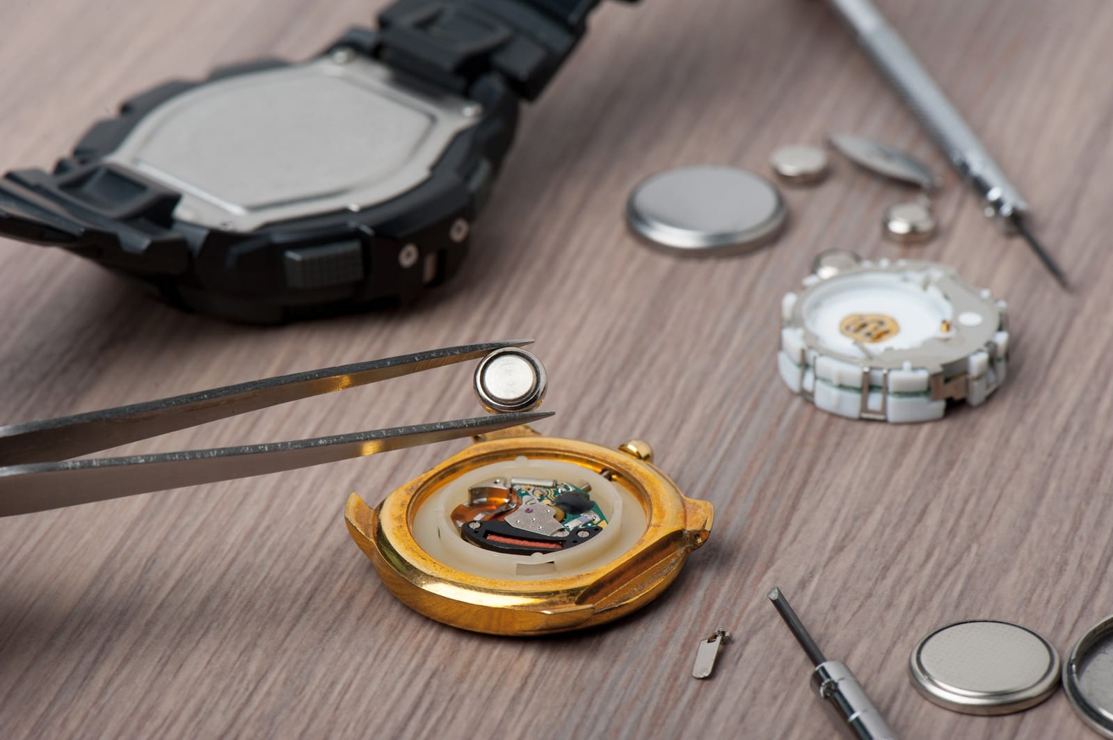 Orologio digitale aperto con pinzetta, batteria a bottone e componenti su un banco da lavoro